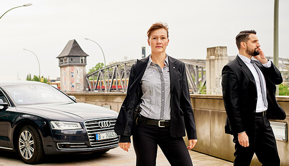 Eine BKA Kriminalkommissarin und ihr telefonierender Kollege stehen in einem Anzug vor einem Auto auf einer Brücke.